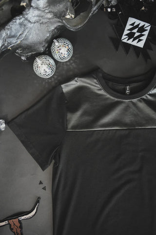 A *MIDNIGHT* LONESTAR SLATE t-shirt from Western Edge Boutique, featuring a luxurious leather-like upper panel, is laid out on a dark surface. Above it, two small disco balls and an abstract geometric decoration with black and white triangles are placed. A small patch with a horned animal design is partially visible in the bottom left.