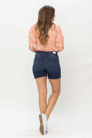 A woman with long, wavy hair stands facing away. She is wearing a short-sleeve, orange and white gingham blouse and Judy Blue Kasey Mid Rise Mid Length Cut Offs Shorts. She completes the casual edgy look with white sneakers. The background is plain white.