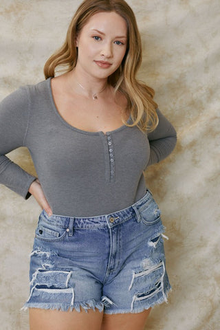 A person with long hair wears a long-sleeved, gray textured top featuring buttons along the front and Sadie Curvy High Rise Mom Shorts from Western Edge Boutique, made of 100% cotton with a 4" inseam. They are posing against a neutral, textured background with a relaxed expression and one hand on their hip.