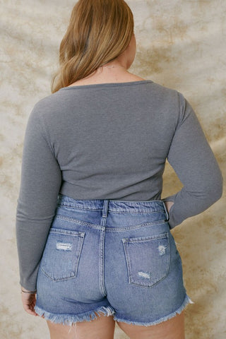 A person with long, light brown hair is facing away from the camera, wearing a gray long-sleeved shirt and Western Edge Boutique's Sadie Curvy High Rise Mom Shorts. The 100% cotton shorts feature a 4" inseam, frayed edges, and distressed details on the pockets. The background is a neutral, textured fabric.