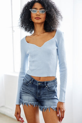 A person with curly hair is wearing cat-eye sunglasses, a light blue long-sleeve ribbed top with a sweetheart neckline, and the Kashlyn Mid Rise Stretch Denim Shorts from Western Edge Boutique. They are posing in a well-lit room with white walls, showcasing a perfect summer look.