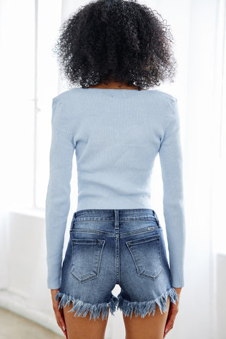 A person with curly hair stands facing away from the camera, wearing a long-sleeved light blue ribbed top and Western Edge Boutique's Kashlyn Mid Rise Stretch Denim Shorts with frayed hems. The person is in a well-lit room with large windows, capturing the perfect summer look.