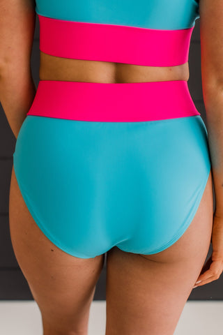 A person is wearing the Ray Of Sunshine Swim Bottoms - Blue & Bright Pink from Western Edge Boutique, part of an exquisite swimwear collection. The two-piece swimsuit features a high-waisted bottom and coordinating top with wide pink bands in vibrant turquoise and bright pink hues. Designed for a comfortable fit, the background remains beautifully out of focus.