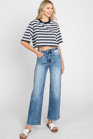 A woman with blonde hair is smiling and wearing a black and white striped crop top paired with the Kennedy High Rise Stretch Classic Straight Jeans from Petra153. These stretchy jeans feature a wide-leg cut and classic 5-pocket styling. She completes her look with white slide sandals, standing confidently against a plain white background, her left hand resting on her hip.