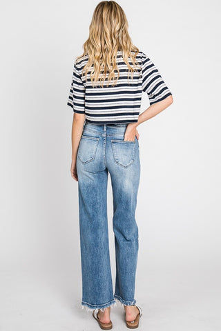 A person with wavy, blonde hair is standing with their back to the viewer. They are wearing a striped black and white short-sleeved shirt, Kennedy High Rise Stretch Classic Straight Jeans from Petra153 that feature a 5-pocket styling and a slightly frayed hem at the bottom, and sandals.