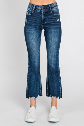 A person is wearing Petra153's Josslyn High Rise Crop Bootcut Frayed Hem Jeans in dark blue, featuring slight distressing. They are also wearing white sneakers. The top and feet of the person are not visible against a plain white background.