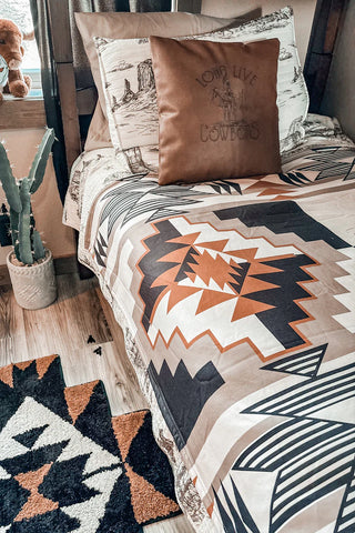 A SANTA FE DUST Blanket from Western Edge Boutique, featuring a southwestern pattern, is draped across the bed with matching pillows near a window adorned with a small dinosaur toy. On the wooden floor, a potted cactus sits alongside a modern Aztec rug, creating a cozy and rustic room atmosphere.