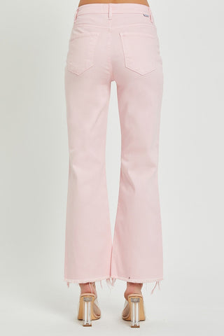 A person wearing Pixley Baby Pink High Rise Straight Crop Pants Distressed by Risen stands with their back facing the camera. The jeans feature distressed knees, two back pockets, and frayed hems, paired with clear high-heeled shoes. The background is plain white.