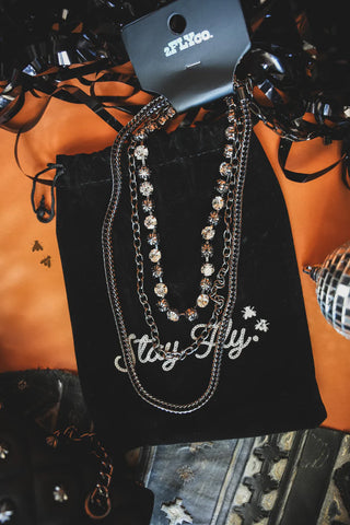 The Midnight Fly Necklace Set, a collection of silver chain necklaces with some featuring decorative beads, is showcased on a black drawstring bag embroidered with "Missy Aly" in white. The display also includes a black ribbon and glimpses of other patterned and studded accessories against an orange backdrop, creating an edgy yet high-quality atmosphere—all under the 2 Fly brand.