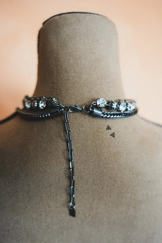 A close-up view of a mannequin's neck featuring the 2 Fly Midnight Fly Necklace Set, adorned with large, clear gemstones. The necklace boasts a quality black chain, delicately hanging down the back and hooked to an adjustable clasp. The softly blurred background in warm tones adds an edgy flair to the overall aesthetic.