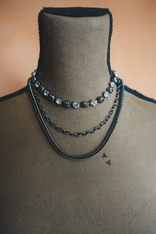 The Midnight Fly Necklace Set by 2 Fly is displayed on a mannequin bust, showcasing three layered necklaces. The top necklace, adorned with sparkling silver beads, exudes quality and elegance. The middle piece features a simple silver chain, while the bottom necklace boasts an edgy, thicker woven silver chain. The warm, muted orange background enhances the overall presentation.