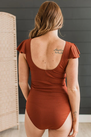 A person with long, wavy hair is facing away, showcasing the MACK & MAI Rusty One Piece Swimsuit with Ruffle Sleeve. The swimsuit is a rust-colored, short-sleeve design featuring a scoop-back and made of ribbed knit material. A tattoo adorns their upper back and another graces their forearm. The scene's backdrop includes a dark paneled wall and a woven room divider.