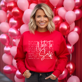 A smiling woman amidst pink and red balloons wears a Western Edge Boutique Amara Note to Self Red Sweatshirt featuring an inspiring quote. With short blonde hair and her hand in her pocket, she exudes confidence and charm in this cozy empowerment setting.