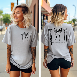 A woman with short blonde hair is wearing Western Edge Boutique's "She Bows Down for No One" tee, strolling down a city sidewalk. The graphic tee is adorned with black bow designs and inspiring text on the back, as she smiles amidst buildings and trees in the background.