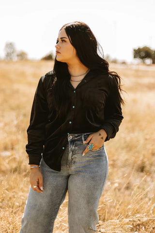 A woman with long dark hair stands in a sunlit field wearing the Saddle Up Velvet Moves top and jeans from Western Edge Boutique. She has turquoise jewelry adorning her hand and wrist, exuding leather lux vibes as she gazes to the side, with tall grasses and a few trees framing the background.