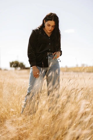 A person with long hair stands in a field of tall, golden grass under a clear sky. They are wearing the versatile Saddle Up Velvet Moves from Western Edge Boutique paired with blue jeans, looking down thoughtfully. Trees and the distant landscape are visible in the background.