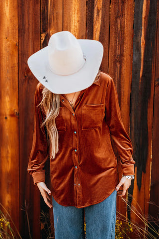 A person in a white cowboy hat is seen looking down, sporting the Saddle Up Velvet Moves shirt from Western Edge Boutique and jeans. They stand before a wooden fence, their face partly hidden, emanating leather luxe vibes with rustic charm.