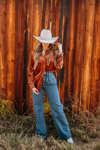 A person in a white cowboy hat, wearing the "Saddle Up Velvet Moves" blouse from Western Edge Boutique, paired with blue jeans, poses in front of a wooden fence. Grass and yellow wildflowers flourish at the base of the fence, adding to the scene's rustic charm.