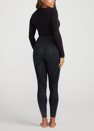 A person is standing barefoot, facing away from the camera. They are wearing a long-sleeved black top and the Elektra Coated Shaping Leggings in black by Yummie, featuring seamless construction. The background is plain white.