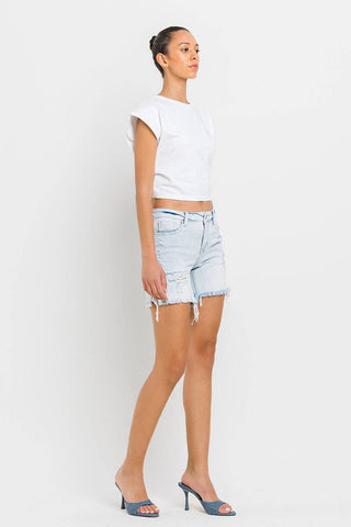 A person with dark hair in a bun is wearing a white sleeveless crop top, Kolson Mid Rise Distressed Stretch Denim Shorts by VERVET by FLYING MONKEY in light blue denim, and blue high-heeled sandals. They are standing sideways against a plain white background.