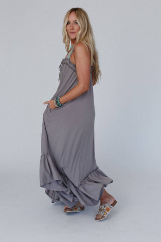 A woman with long blond hair is standing in profile, wearing the Three Bird Nest Heavenly Glow Maxi Dress in Mushroom with ruffled details at the hem. She accessorizes her boho style with turquoise bracelets and brown sandals adorned with white patterns, smiling at the camera against a plain white background.