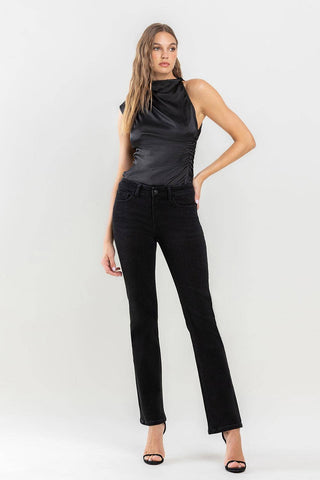 A woman with long hair is posing against a plain white background, showcasing a sleek black sleeveless top and Halston Mid Rise Bootcut Jeans by Lovervet by VERVET. She completes the look with black high-heeled sandals, creating a true wardrobe must-have. With one hand on her hip and a confident stance, she exudes style.