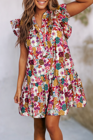 A woman with wavy hair smiles and poses with one arm bent and raised. She is wearing a Kace Floral Printed Split Neck Flutter Sleeve Dress from Little Daisy Closet. The lightweight blend dress features a vibrant pattern with various flowers in colors such as red, pink, yellow, and green—perfect for your summer wardrobe. The background is blurred.