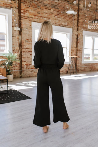 A person with blonde hair stands barefoot in a bright, airy room with large windows and exposed brick walls. They are facing away from the camera, dressed in the Alex Black Lounge Set by LIAM AND CO—featuring a versatile black long-sleeve top and matching wide-leg pants that balance comfort and style.