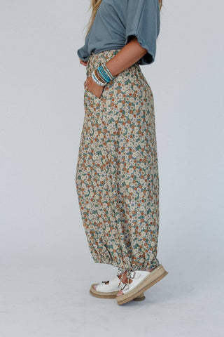 A person wearing a light blue top with elbow-length sleeves, the Solstice Song Cinched Skirt in Khaki from Three Bird Nest featuring a boho mix of small flowers in green, white, and pink, and white platform sandals. The person has their hands in their pockets and is accessorized with multiple stackable bracelets.