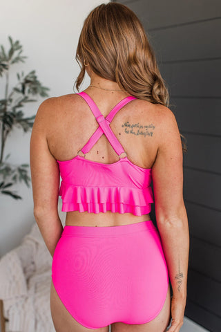 The image portrays the back view of an individual with long hair adorned in the Island Essence Hot Pink Swim Top by Western Edge Boutique. The swimsuit is highlighted by its X-strap back and adjustable straps. Additionally, the person has a tattoo on their shoulder, with a plant and light-colored furniture appearing in the background.