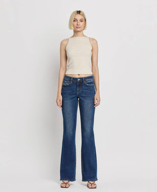 A woman stands against a plain white background wearing a beige sleeveless top and Kip Mid Rise Relaxed Bootcut Jeans by Lovervet by VERVET. She has short blond hair and is wearing brown sandals. She is facing the camera with a neutral expression.