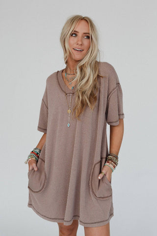 A woman with long, wavy blonde hair wears the Brooklyn Soft Touch Tunic Dress by Three Bird Nest, a loose, short-sleeve dress made of comfortable stretch fabric in a taupe boho-chic style. She accessorizes with layered necklaces and colorful bracelets on both wrists. She has a relaxed expression and stands against a plain white background.