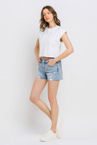 A person with long, wavy hair is posing against a plain white background. They are wearing a white short-sleeve crop top, VERVET by FLYING MONKEY's Kylie Mid Rise Raw Distressed Hem shorts in comfortable stretch denim, and white sneakers. They have their left hand in their pocket and are looking forward with a relaxed expression.