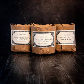 Three bars of soap wrapped in brown paper sit on a dark surface. Each soap has a white label with ornate black borders, featuring the names: "Rose Rebellion," "Cadillac Cowgirl," and "Sweet Rebellion." Alongside them rests an 8 oz. Harvest Moon Candle from R. Rebellion, made of all-natural coconut soy wax with wood wicks, enhancing the rustic feel.