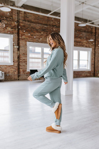 A woman with long hair, happily walking indoors in a spacious, well-lit room with large windows and exposed brick walls, is wearing tan furry boots and the Alison Half Zip Loungewear Set by Liam & Company. She is smiling and appears to be relaxed in her cozy two-piece outfit.