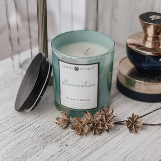 A green Illumination Candle from Jewel Within, labeled "Valencia Orange" and crafted from natural coconut wax, is displayed on a white wooden surface. The candle's black lid lies next to it. Decorative metallic items provide a stylish backdrop, while dried star-shaped flowers are arranged in front of the candle.