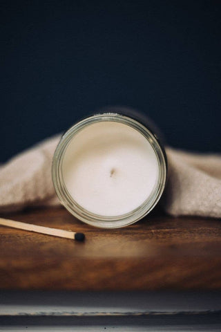 A Luxe(Diva) Candle in a white, 8 oz glass jar from Mav Wicks Candle Co rests on a wooden surface with a single matchstick lying next to it. A beige cloth peeks out from beneath the jar. The dark background provides contrast to the candle and its surroundings, creating an inviting home decor accent.