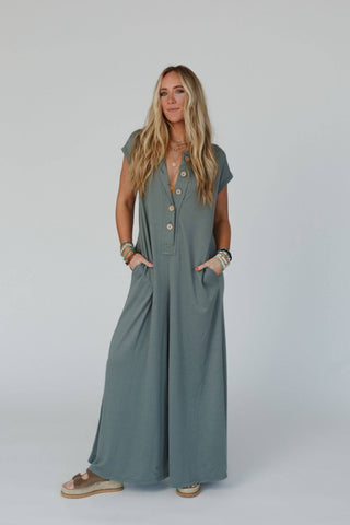 Against a plain background, a woman with long, blonde hair confidently poses wearing the Logan Wide Leg Jumpsuit in light olive from Three Bird Nest. This loose-fitting, short-sleeved jumpsuit features buttons down the front, side pockets, and is made from comfortable knit fabric. It pairs perfectly with her bracelets, necklaces, and sandals.