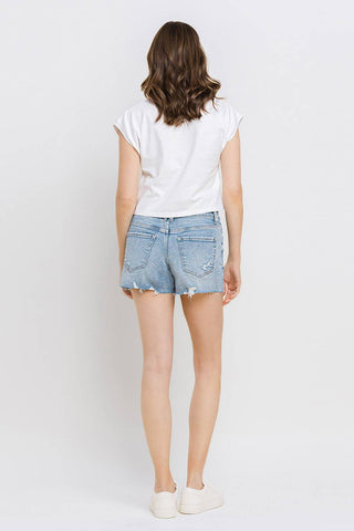 A person with shoulder-length brown hair is standing with their back to the camera, wearing a white short-sleeve top, Kylie Mid Rise Raw Distressed Hem shorts in comfortable stretch denim from VERVET by FLYING MONKEY, and white sneakers. The background is a plain white wall. Available in all sizes.