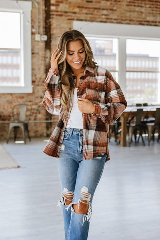 A person with long hair smiles while standing indoors, wearing the Kelsea Plaid Shacket by Liam & Company over a white top and distressed blue jeans. The scene is set against a brick wall, with large windows and a wooden table with chairs completing the cozy ambiance.