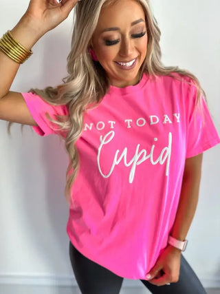 A woman with long blonde hair wears the Bird & Co Wholesale Tees' "Not Today Cupid" neon pink graphic tee, paired with black pants and gold bracelets, while gently holding a hair strand and exuding confidence.