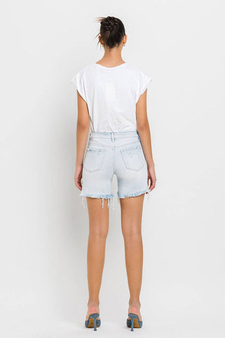 A person stands facing away from the camera, wearing a white T-shirt and Kolson Mid Rise Distressed Stretch Denim Shorts by VERVET by FLYING MONKEY. They are also sporting stylish blue and tan high-heeled shoes. Their hair is tied up in a casual bun. The background is plain white, creating a minimalist look.