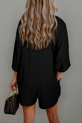 A person with long, wavy blonde hair is standing facing away, wearing a chic black Brecken Women Half Sleeve Button Collared Loose Fit Romper by Aapparella and holding a black handbag with a chain strap in their right hand. The background is plain and light-colored, perfect for highlighting elegant pieces in women's fashion.