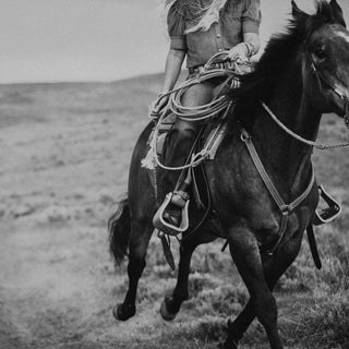 A black and white image captures the timeless elegance of musk, featuring a person riding a horse along a grassy trail. The rider, dressed casually, holds a coiled rope while the horse moves energetically through the vast, open landscape. This scene evokes the essence of Western Skies Roll On Perfume Oil by R. Rebellion.