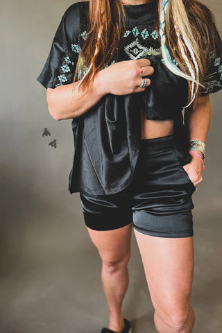 A person wearing a black shirt with an embroidered design and Layken Cowboy Tuxedo Shorts by 2 Fly stands against a neutral background. Their face is not visible. They have long hair with a streak of blonde, and they are adorned with multiple bracelets and rings. The shorts feature a discreet side zipper for added style.