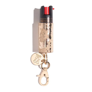 The BLINGSTING Emergency Pepper Spray Key Ring boasts a gold-colored canister adorned with a marble pattern. It comes equipped with a carabiner clip and features a round charm engraved with the brand's name, "Blingsting," from the renowned women's safety brand. The black trigger mechanism includes a red button and UV marking dye, all wrapped up in a compact and stylish design.