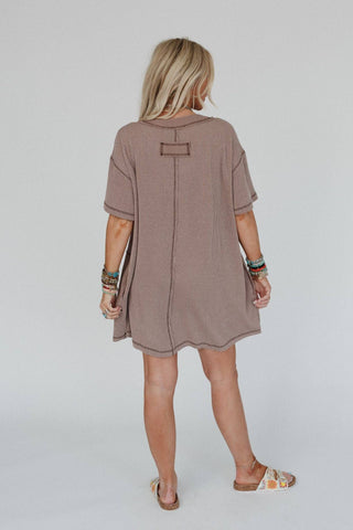 A woman with long blond hair stands facing away from the camera, wearing a boho-chic Brooklyn Soft Touch Tunic Dress by Three Bird Nest in a loose, light brown short-sleeved style. She accessorizes with colorful bracelets on both wrists and floral-patterned slides, standing against a plain white background.