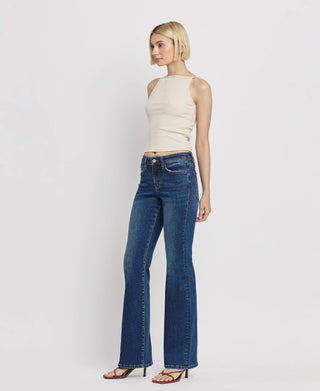 A woman with short, blonde hair stands against a plain white background. She is wearing a sleeveless, beige top and Lovervet by VERVET's Kip Mid Rise Relaxed Bootcut Jeans in dark blue. Complementing her outfit are black strappy heels.