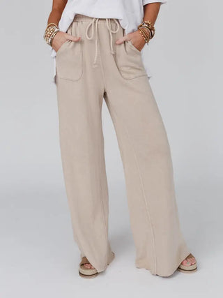 Person wearing loose, wide-leg Cooper Relaxing Robin Pants in light beige with a drawstring waist and large patch pockets featuring a vintage fade effect, paired with a white top and multiple beaded bracelets. The outfit exudes a casual and relaxed vibe, with the pants slightly covering their footwear. The background is plain light grey.