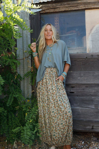 A woman with long blonde hair stands in front of a rustic wooden structure amidst lush greenery. She is wearing a loose-fitting gray top paired with the Three Bird Nest Solstice Song Cinched Skirt in Khaki, accessorized with various bracelets and necklaces. With one hand in her skirt pocket, she leans against the wall.